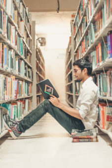 Young man sitting on floor of library reading a book.